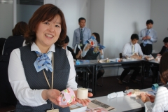 ＥＭ石鹸と梱包された完成品を手に持つ平野靖子さんと、梱包作業に勤しむJA共済役職員